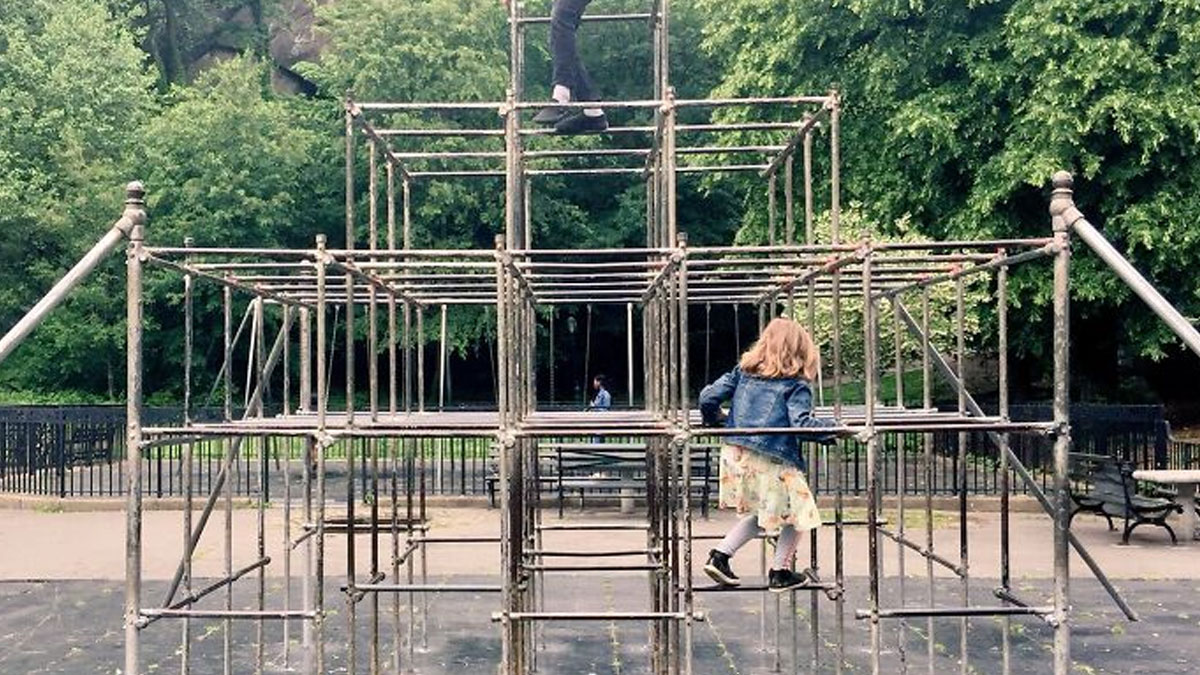 Child climbing on a historic metal playground structure surrounded by trees, showing old risky playground equipment.