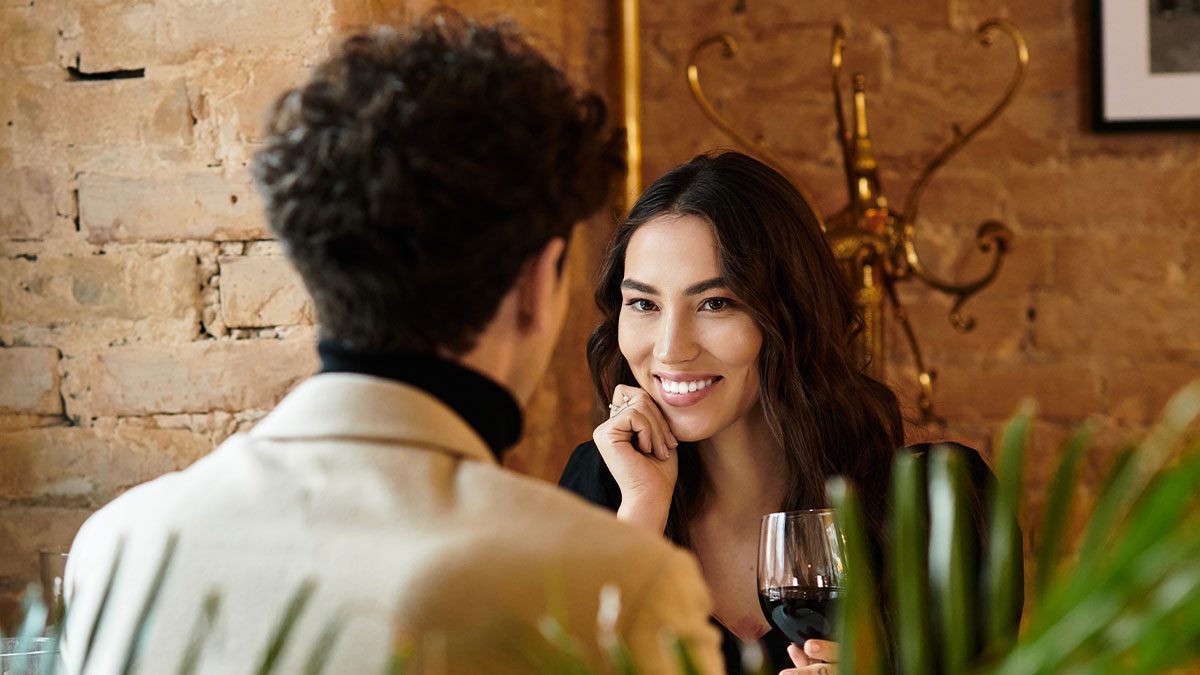Widow and single mom on a date in a cozy restaurant, smiling and holding a glass of red wine during conversation.