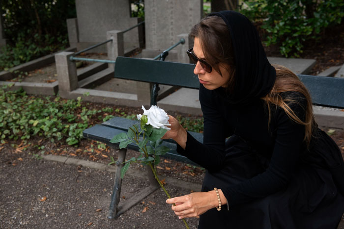 Widow wearing black and sunglasses sits on park bench holding a white rose, reflecting with sadness after family dinner conflict.