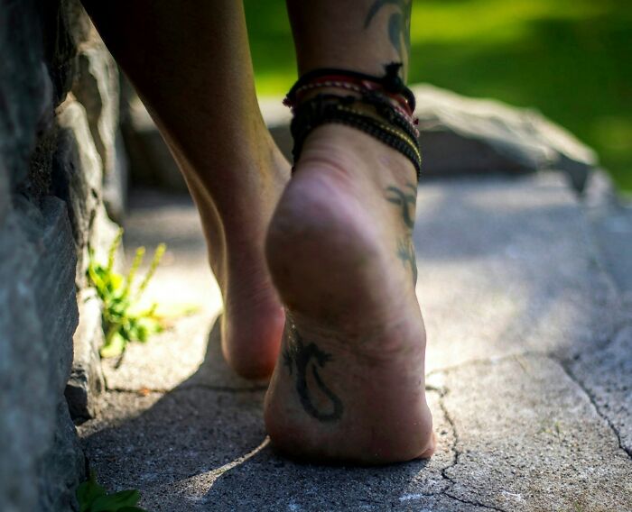 Close-up of a bare foot with multiple tattoos and bracelets walking on a stone path, highlighting tattoo art details.