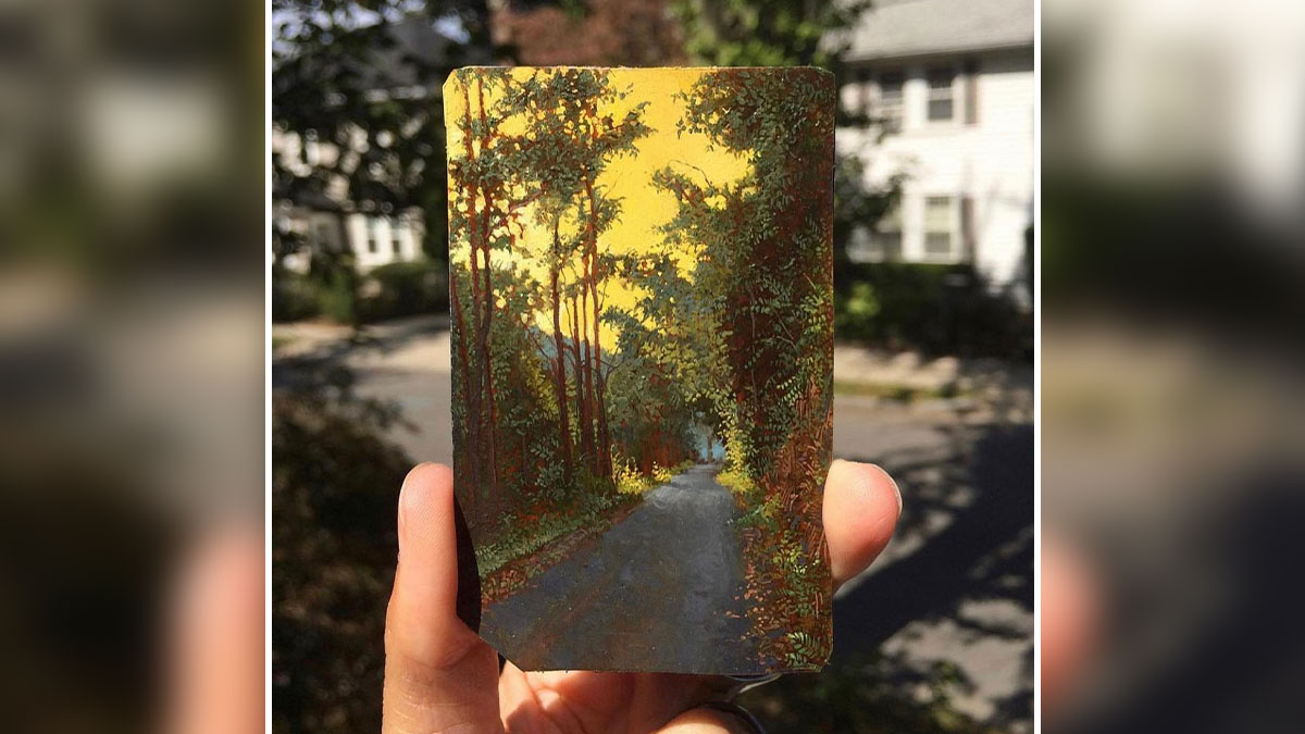 Hand holding a detailed painting by Dina Brodsky showing a tree-lined road with vibrant natural colors.