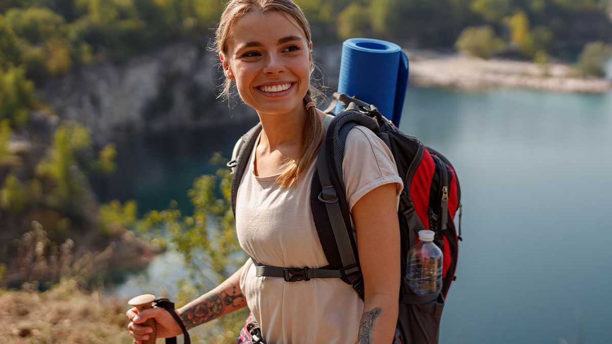 Woman hiking solo with backpack and walking stick, smiling outdoors near a lake and forest landscape.