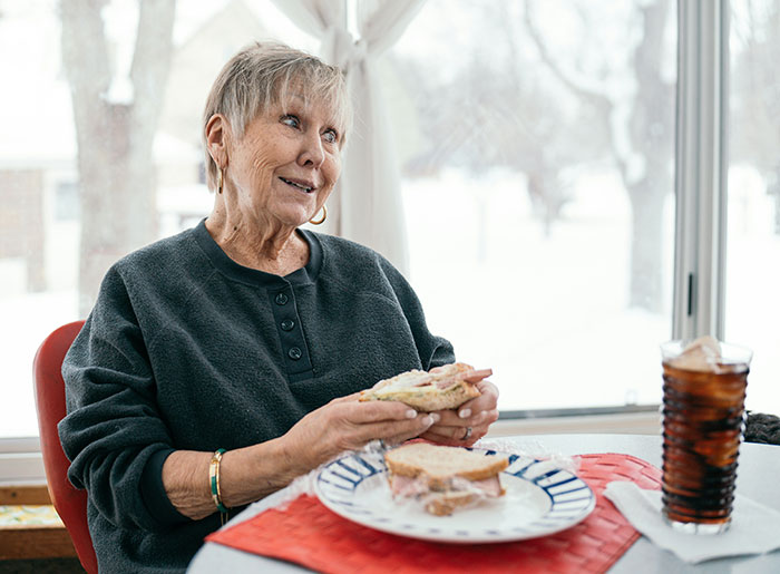 Elderly woman eating sandwich indoors by window, illustrating food that increases dementia risk after long-term study. Elderly woman eating sandwich indoors by window, illustrating food that increases dementia risk after long-term study.