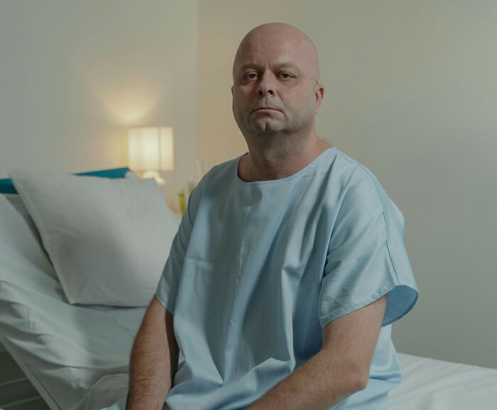 Bald man in hospital gown sitting on bed, representing cancer diagnosis and betrayal from hidden truth. Bald man in hospital gown sitting on bed, representing cancer diagnosis and betrayal from hidden truth.