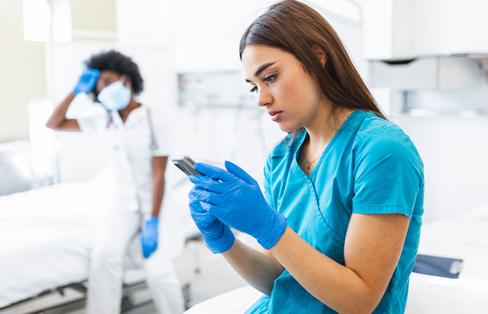 Healthcare workers in blue scrubs and gloves using a phone with a colleague wearing a mask in a medical setting.