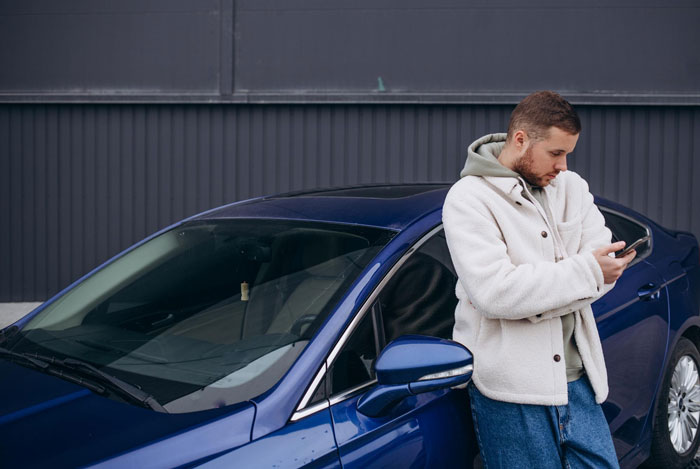 Young man leaning on blue car, checking phone outside a garage, depicting entitled neighbor trying to hijack driveway. Young man leaning on blue car, checking phone outside a garage, depicting entitled neighbor trying to hijack driveway.