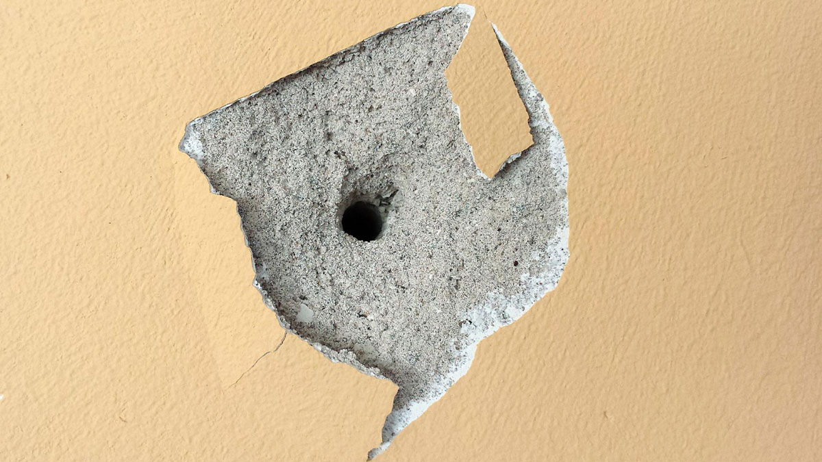 Close-up of nasty damage on beige wall with a large hole and broken plaster revealing rough texture beneath.