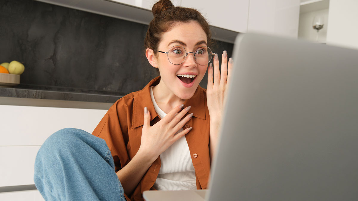 Young woman with glasses excitedly discovering unbelievable facts while looking at a laptop screen in a modern kitchen.