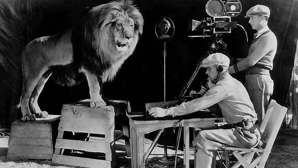 Black and white historical photo of a lion on crates being filmed by two men with vintage camera equipment.