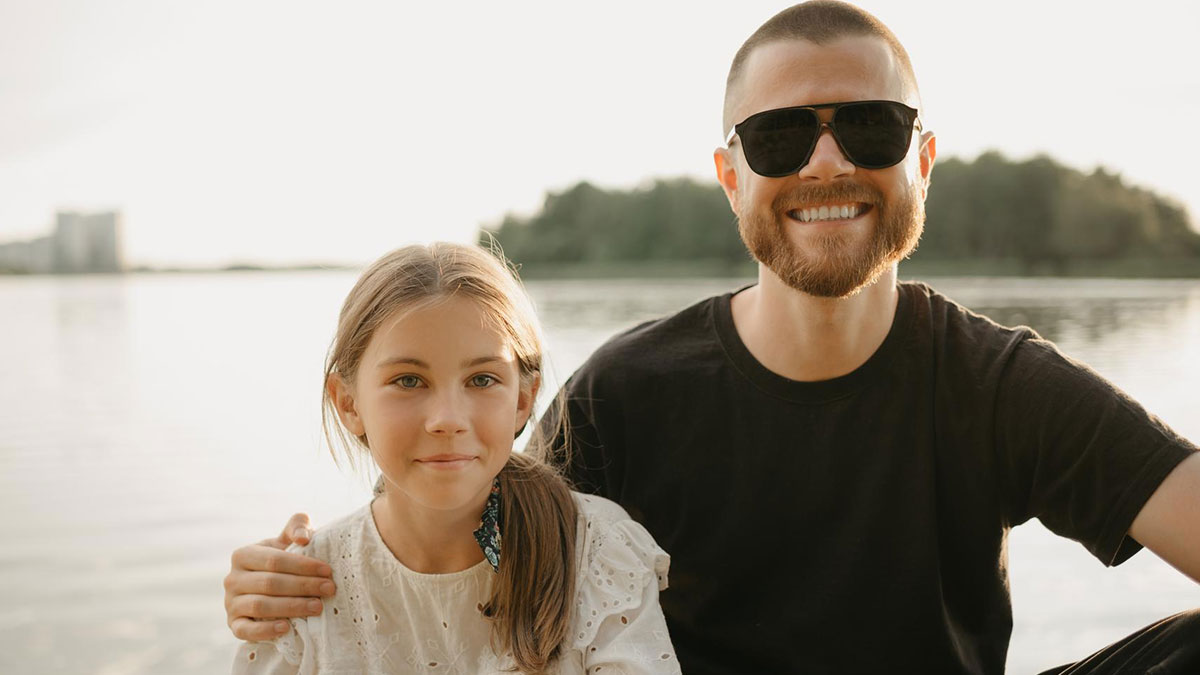 Smiling dad wearing sunglasses with arm around daughter by the lake, illustrating child support and parenting control themes.