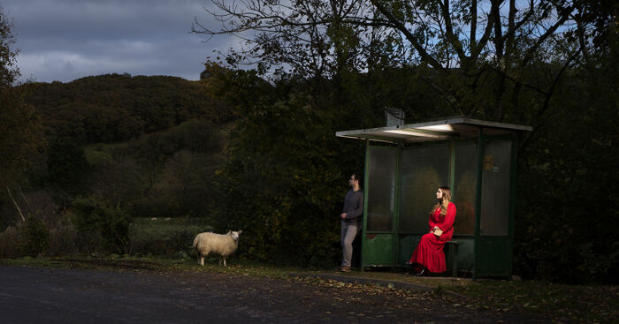 A woman in a red dress and a man stand at a dark bus stop in a rural area, illustrating dying to be heard concept.