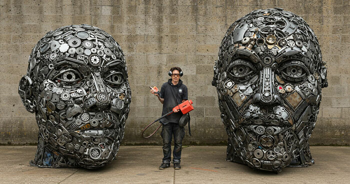 Artist standing between two large stunning sculptures made from trash metal parts against a concrete wall.