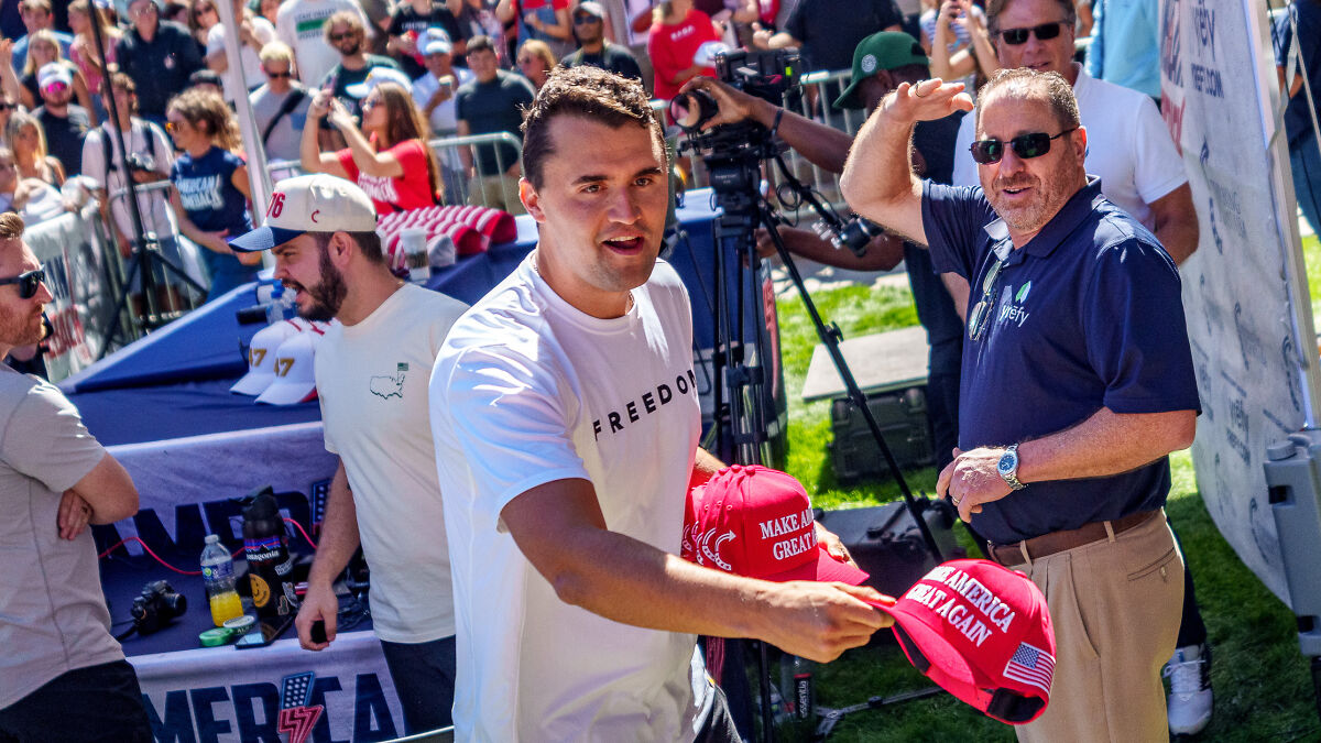 Man in freedom shirt handing out Make America Great Again hats at outdoor event with crowd and cameras behind him