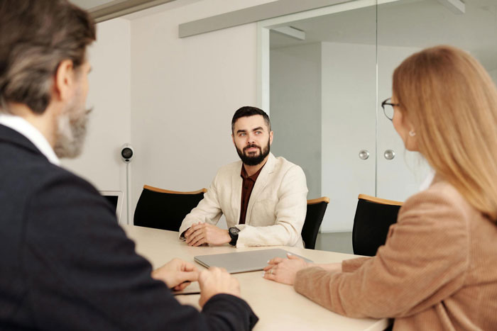 Man in beige jacket sitting at office table with two colleagues during discussion about expensive employee situation