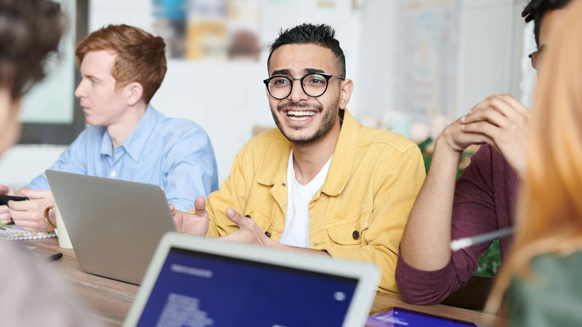 Young man in yellow jacket smiling during a company meeting about an expensive employee with a pricey trick up his sleeve