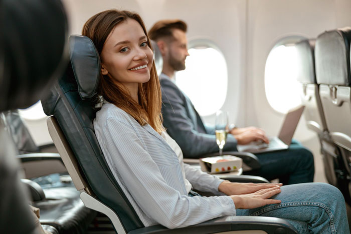 Young woman enjoying her first class flight experience smiling while seated next to a man working on a laptop on a plane. Young woman enjoying her first class flight experience smiling while seated next to a man working on a laptop on a plane.