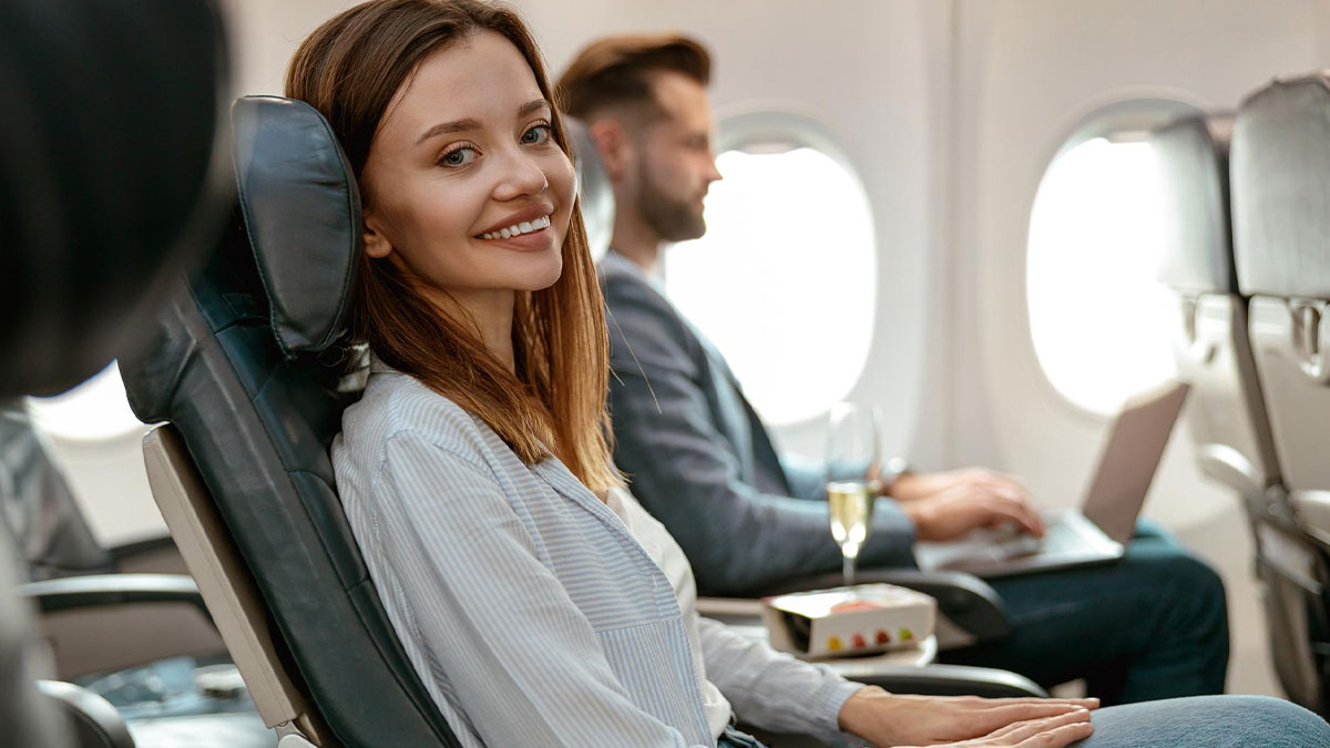 Woman enjoying her first class flight smiling confidently while seated next to a man working on his laptop on a plane.