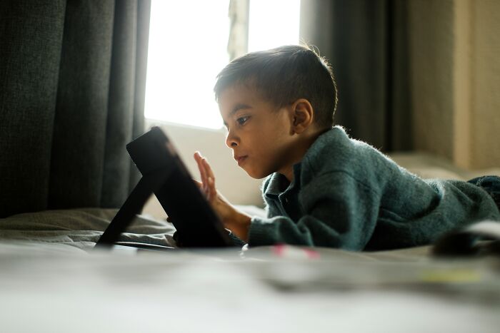 Young boy lying on bed using tablet device focused on screen, illustrating parenting hacks that save sanity.