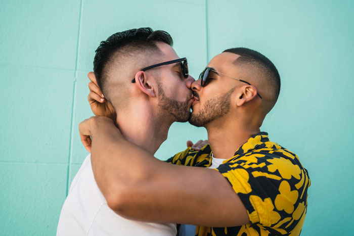 Groom kisses best friend at bachelor party in a close embrace, showing affection against a teal wall background. Groom kisses best friend at bachelor party in a close embrace, showing affection against a teal wall background.