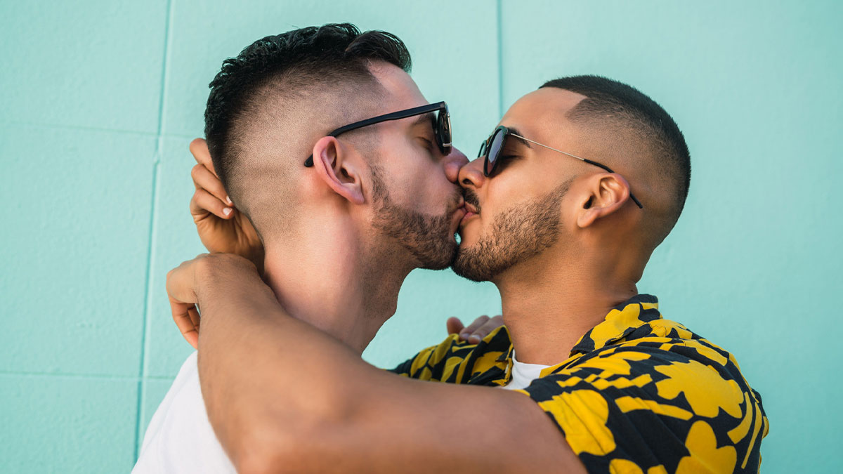 Two men wearing sunglasses share a kiss, symbolizing a groom kissing his best friend at a bachelor party.