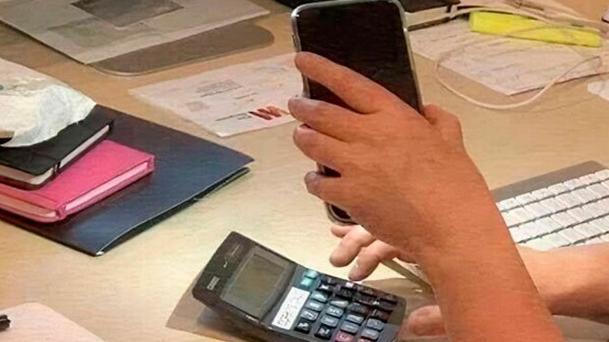 Student using phone with one hand and calculator with the other while sitting at a cluttered desk with papers and notebooks.