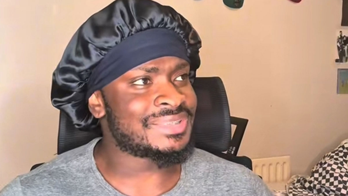 Man wearing a black satin bonnet and gray shirt, sitting in a chair, smiling and looking to the side in a casual room setting.