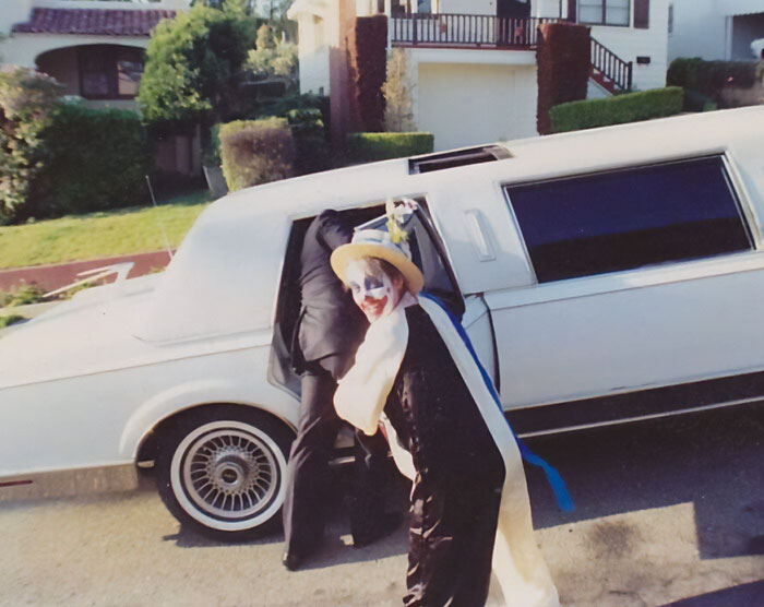 Child dressed as clown smiling near limousine with a parent for a funny parents moment in a suburban neighborhood.