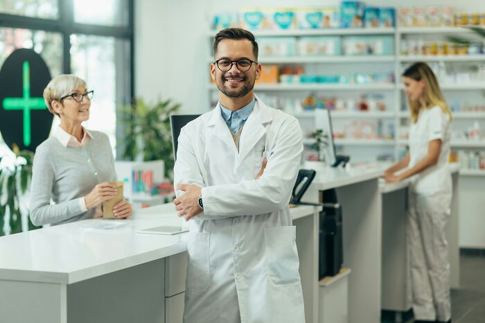 Smiling professional in white coat stands in pharmacy, illustrating suspicious professions like butcher and gravedigger combined.