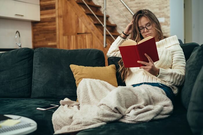 Woman wrapped in blanket on sofa reading a book, reflecting on groom insisting guest attend wedding despite positive Covid test. Woman wrapped in blanket on sofa reading a book, reflecting on groom insisting guest attend wedding despite positive Covid test.
