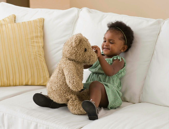 Toddler sitting on white couch playing with teddy bear, illustrating parenting hacks that save sanity.