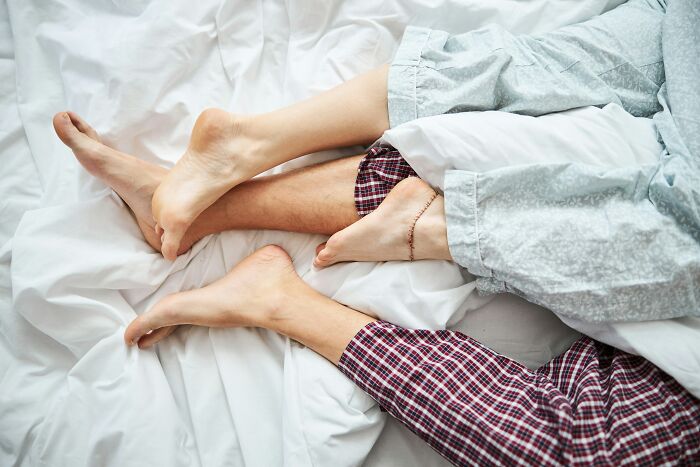 Two people lying closely in bed, feet entangled under covers, depicting intimate moments shared but never admitted.