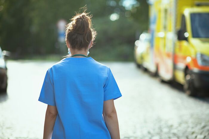 Person in blue scrubs standing outside near blurred emergency vehicles, illustrating professions that raise suspicion when paired.