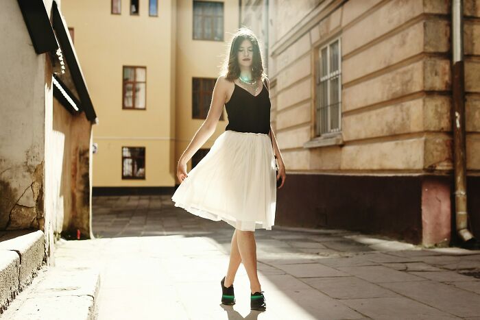 Young woman in a black top and white skirt walking in sunlit urban alley, symbolizing kaomojis cultural expression and creativity.