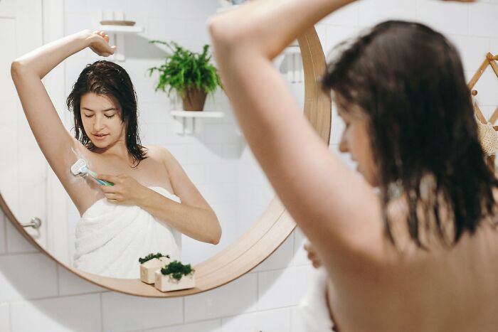 Young woman wrapped in towel shaving her armpit in front of a bathroom mirror, reflecting a casual grooming moment.