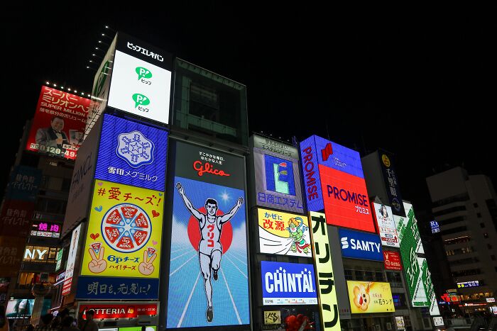 Nighttime cityscape with colorful illuminated billboards, highlighting the contrast of butcher and gravedigger professions.