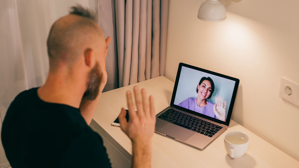 Man in video therapy session on laptop, woman on screen smiling and waving in a private therapy setting.