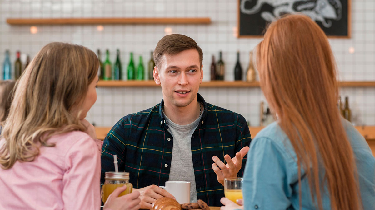 Young man talking to two women at a cafu00e9, discussing a prank to support autistic sister and relationship challenges.