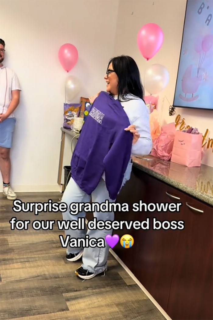 Woman at a surprise grandma baby shower holding a purple sweatshirt, celebrating her new status with balloons and gifts nearby. Woman at a surprise grandma baby shower holding a purple sweatshirt, celebrating her new status with balloons and gifts nearby.