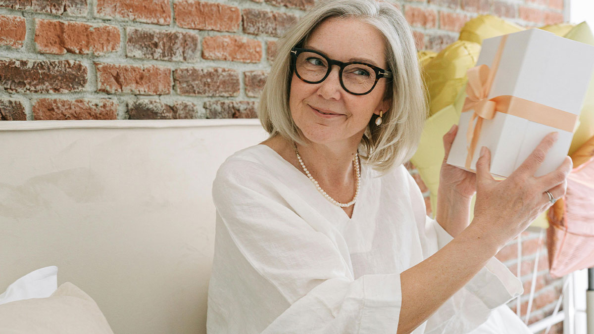 Grandma in glasses holding a gift box, smiling, with balloons in the background at a baby shower celebration.