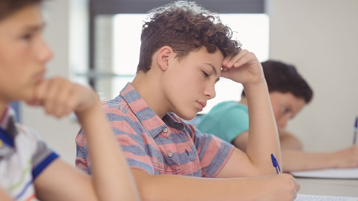 Teen boy focused and stressed while writing in a notebook during a group project with lazy teammates in a classroom setting