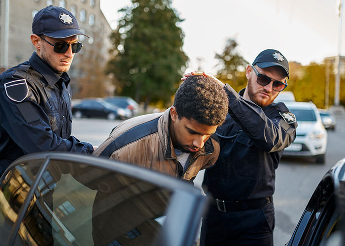 Man being arrested by two police officers outdoors near parked cars on a city street, woman considers canceling holiday booking. Man being arrested by two police officers outdoors near parked cars on a city street, woman considers canceling holiday booking.