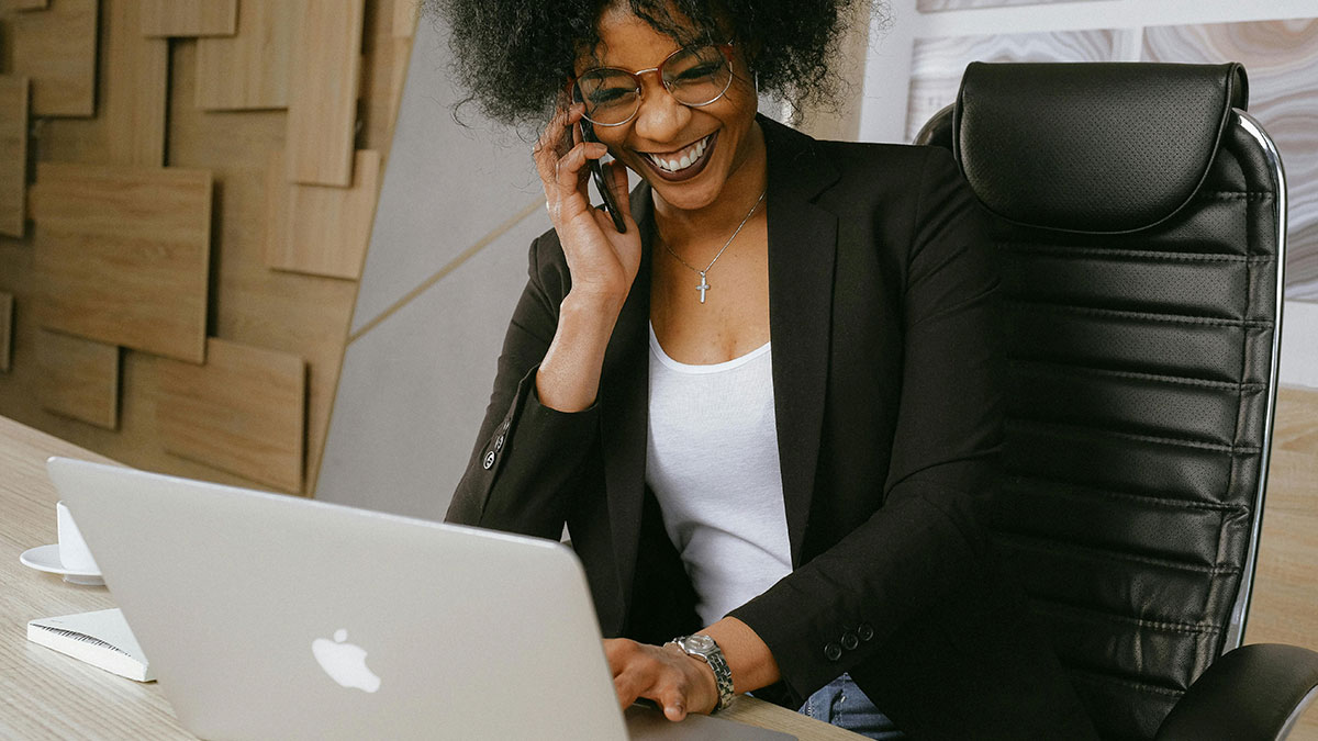 Smiling working mom talking on phone while using laptop in modern office, representing hurt working mom and ignorant husband.