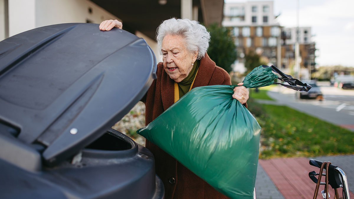 Elderly woman digging through trash with a green bag near a dumpster in a residential area.