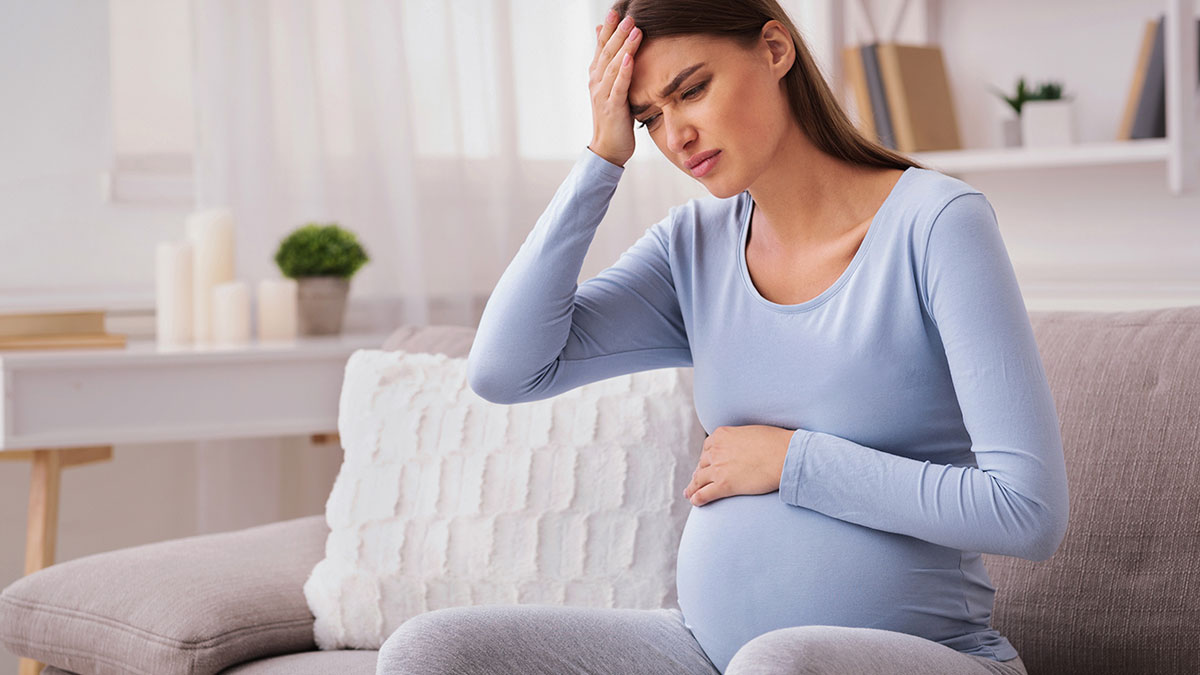 Pregnant woman in blue shirt sitting on couch, holding belly and forehead, showing discomfort and stress at home.