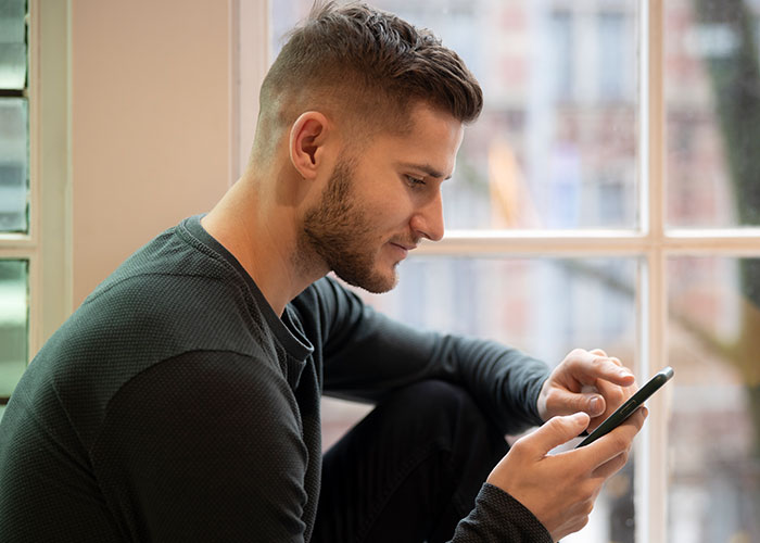 Young man sitting by a window, looking focused and suspicious while checking his smartphone indoors. Young man sitting by a window, looking focused and suspicious while checking his smartphone indoors.