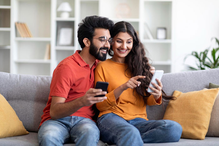 Couple sitting on a couch smiling and looking at phones, discussing husband parents native language at home. Couple sitting on a couch smiling and looking at phones, discussing husband parents native language at home.