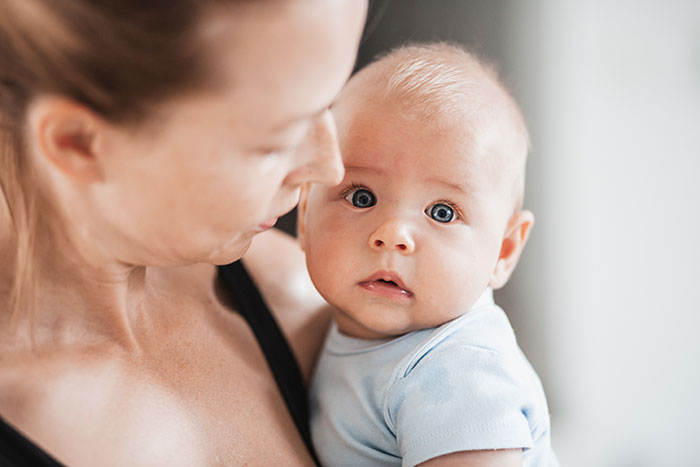 New mom holding her baby with a worried expression, struggling with baby care while husband judging silently nearby. New mom holding her baby with a worried expression, struggling with baby care while husband judging silently nearby.