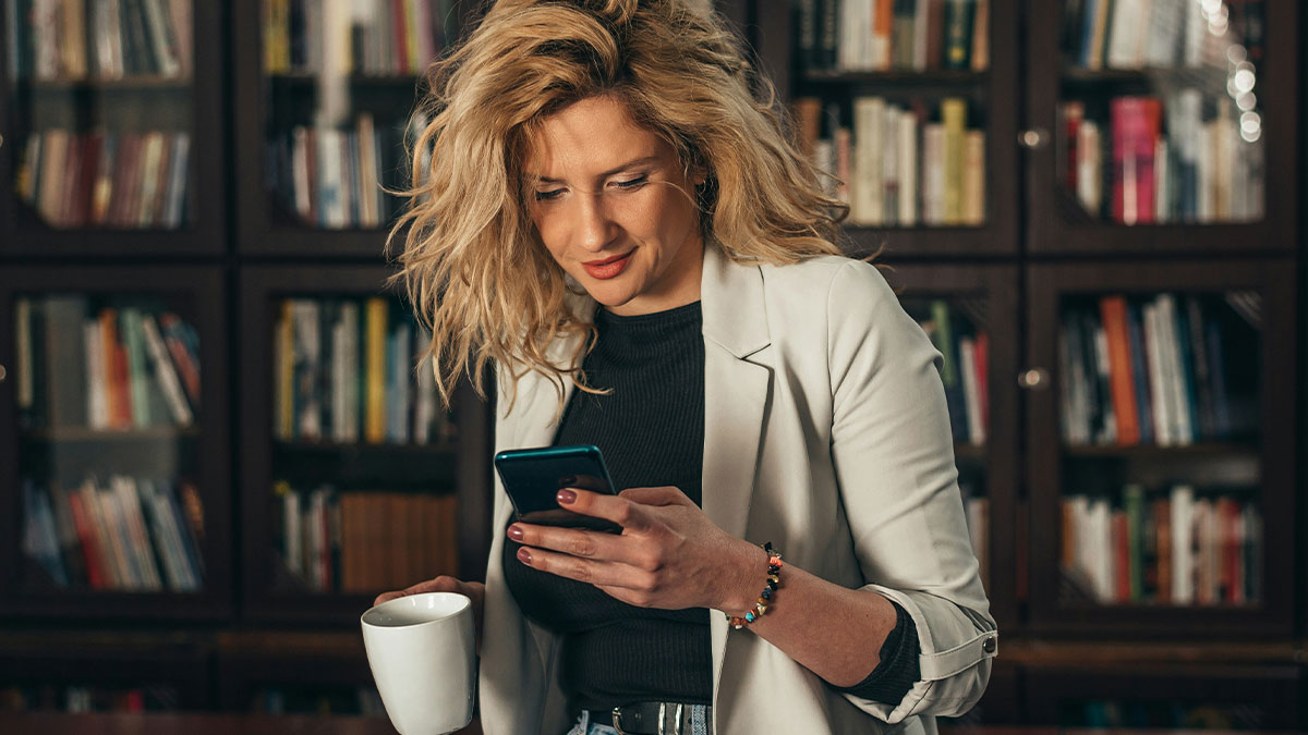 Woman with blonde hair wearing a white blazer reading on phone and holding a coffee mug in a room with bookshelves behind her.