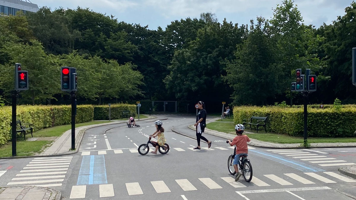 Children riding bikes and an adult walking in a safe Danish prison cell street with traffic lights and greenery.