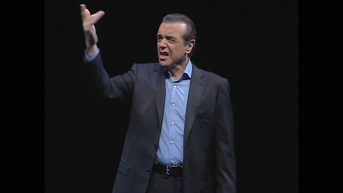 Man in a dark suit and blue shirt gesturing passionately while presenting intriguing facts about daily knowledge on stage.